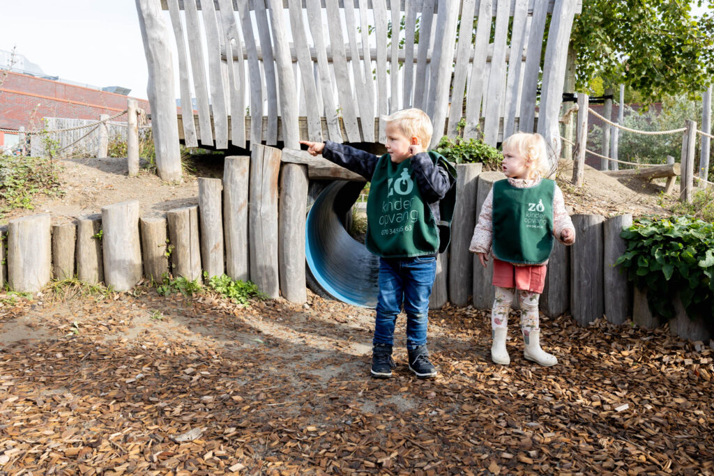 Twee kinderen spelen in de speeltuin. Het jongetje wijst naar iets in de omgeving.