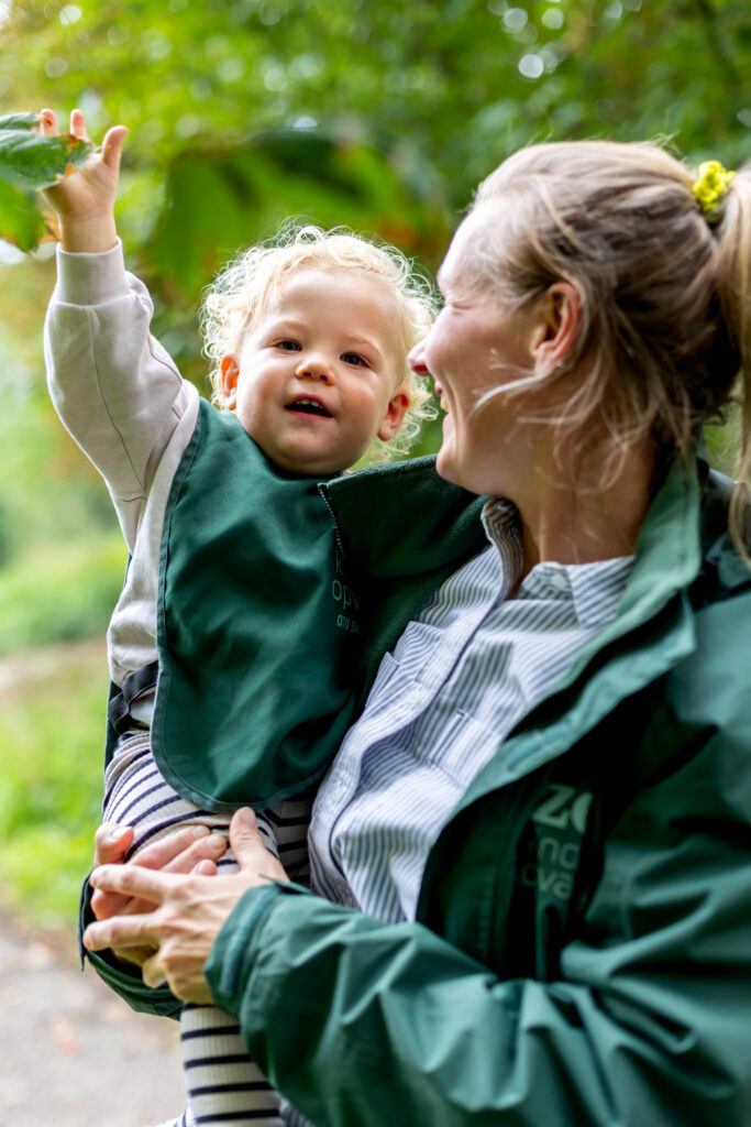 A Zo Kinderopvang childcare practitioner holding a waving toddler in her arms.