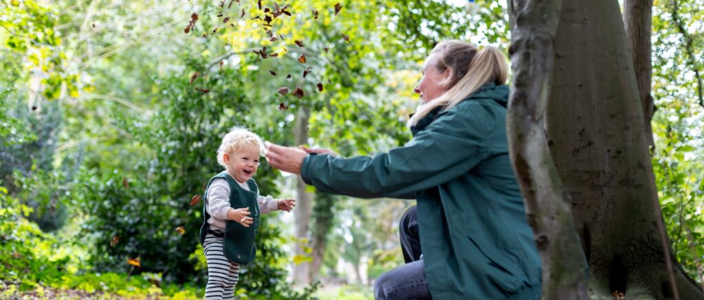 Pedagogisch medewerker en kind staan in het bos en gooien boombladen in de lucht